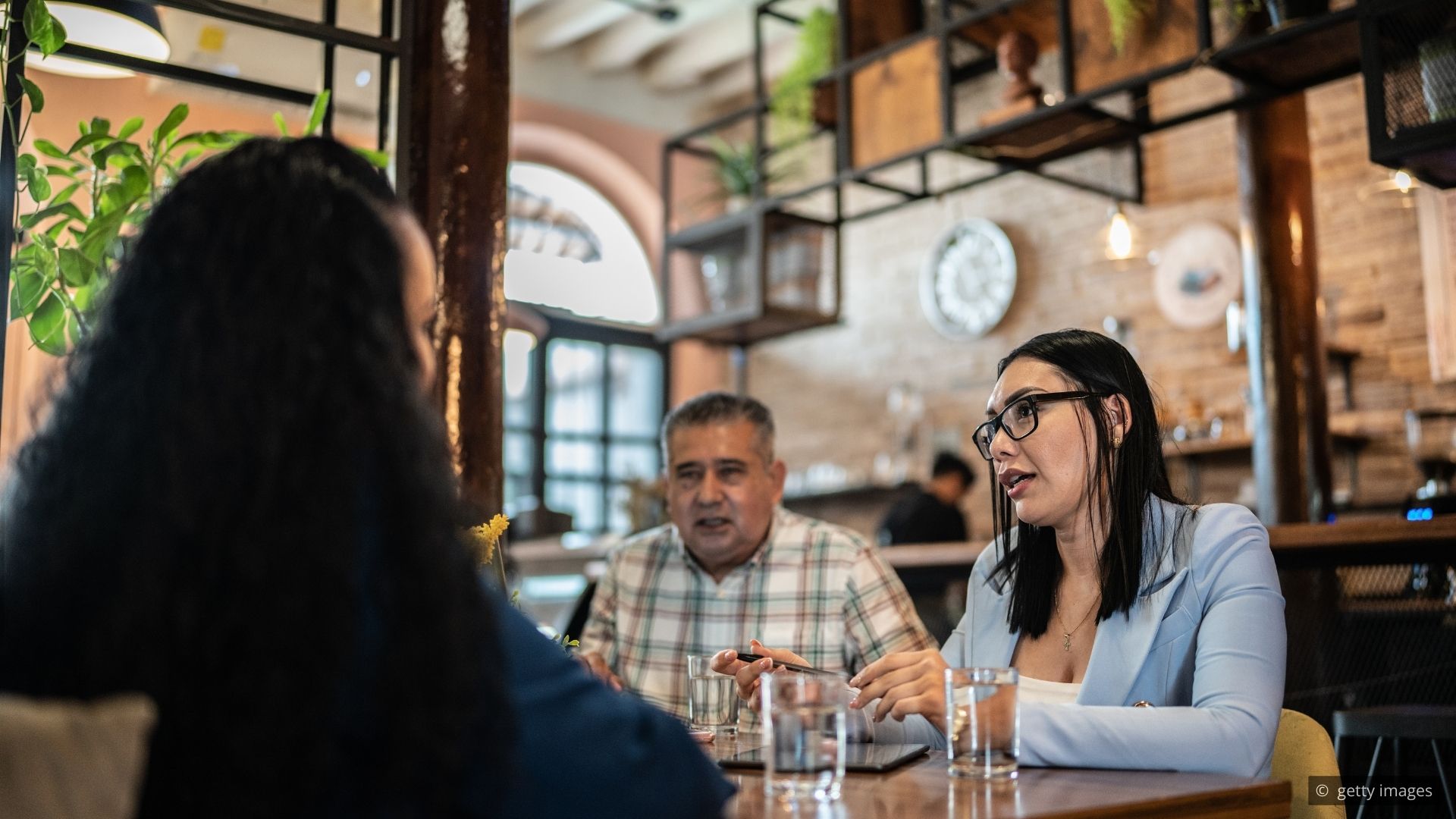 A hotel lounge with a server observing guests from a composed distance before approaching the table